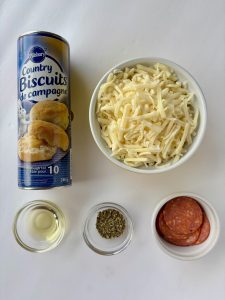 Ingredients used to make pizza bread wreath: a tube of Pillsbury Country Biscuits, a bowl of shredded cheese, a small bowl of oil, a small bowl with seasoning, and a small bowl with pepperoni slices