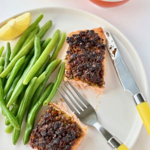 A white plate with a piece of salmon cut in half, green beans, wedge of lemon and yellow cutlery.